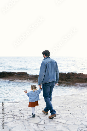 Dad in a denim suit walks by the hand with a little girl in a dress and a denim jacket along the pier against the background of the sea. Back view