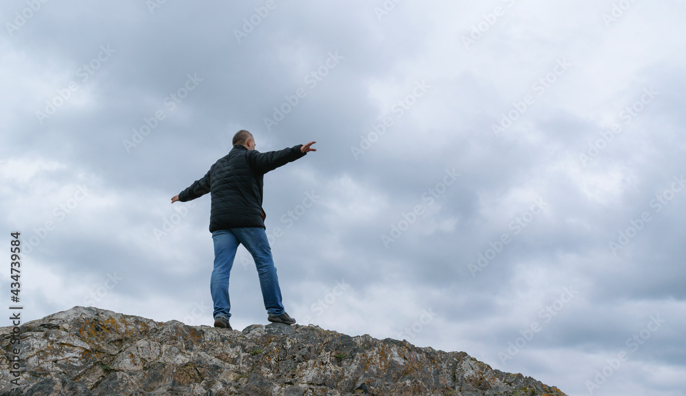 A middle-aged man walks along the ridge of a rock. Hands out to the sides for balance. View from the back. The background is the sky with dramatic clouds. A middle-aged man walks along the ridge of a rock. Hands out to the sides for balance. View from the back. The background is the sky with dramatic clouds.