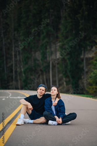 Beautiful young couple spends time together in nature, sitting on the asphalt of a mountain road against the backdrop of a coniferous forest and relaxing.