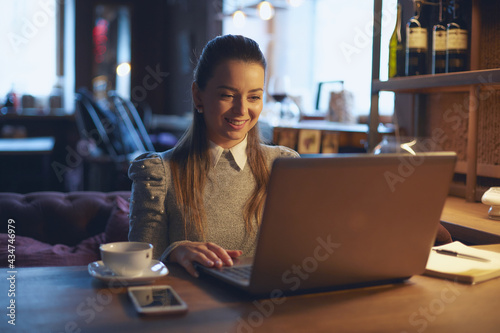Young beautiful girl woman sitting at a wooden table in a cafe indoors  working on a laptop and smiling while communicating with a customer. Remote work and freelance concept.