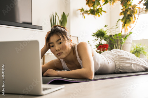 Woman laying at the mat at the floor and watching yoga lesson at the laptop