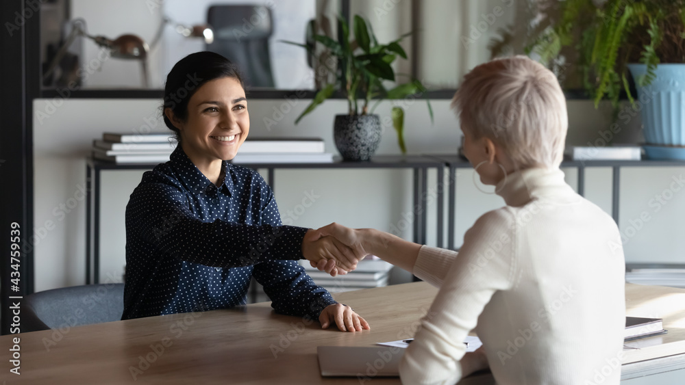 Smiling multiethnic businesswomen shake hands close deal or agreement at meeting in office. Happy young Indian woman handshake business partner get acquainted greet at briefing. Employment concept.
