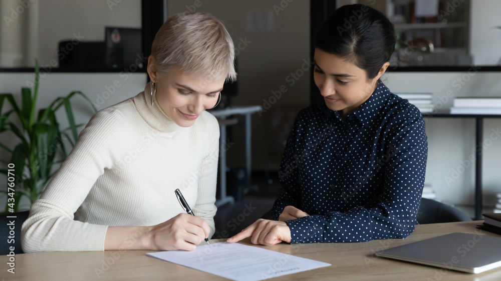 Happy young Caucasian woman sign paper document close deal with Indian business partner or client at meeting. Smiling multiracial businesswomen put signature on paperwork contract make agreement. Happy young Caucasian woman sign paper document close deal with Indian business partner or client at meeting. Smiling multiracial businesswomen put signature on paperwork contract make agreement.