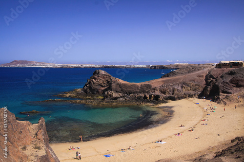 View from steep cliff into secluded blue lagoon sand beach with few people and white village background - Playa Papagayo, Playa Blanca - Lanzarote