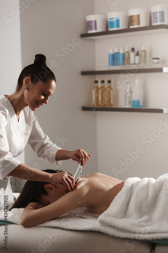 Smiling masseur doing gua sha scrapping tsm treatment. Woman relaxing during gouache massage, lying on therapy table, beautician rubbing client back with asian technique to relieve tension photo