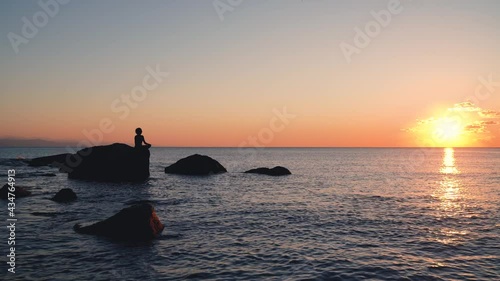 Silhouette of Peaceful woman meditating on a rock in the middle of a low wave sea at sunset time. golden hour colorr photo