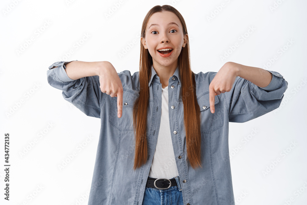 Cheerful young teen female student, pointing fingers down and smiling pleased, demonstrate banner, showing your logo, promotional text on copyspace, white background