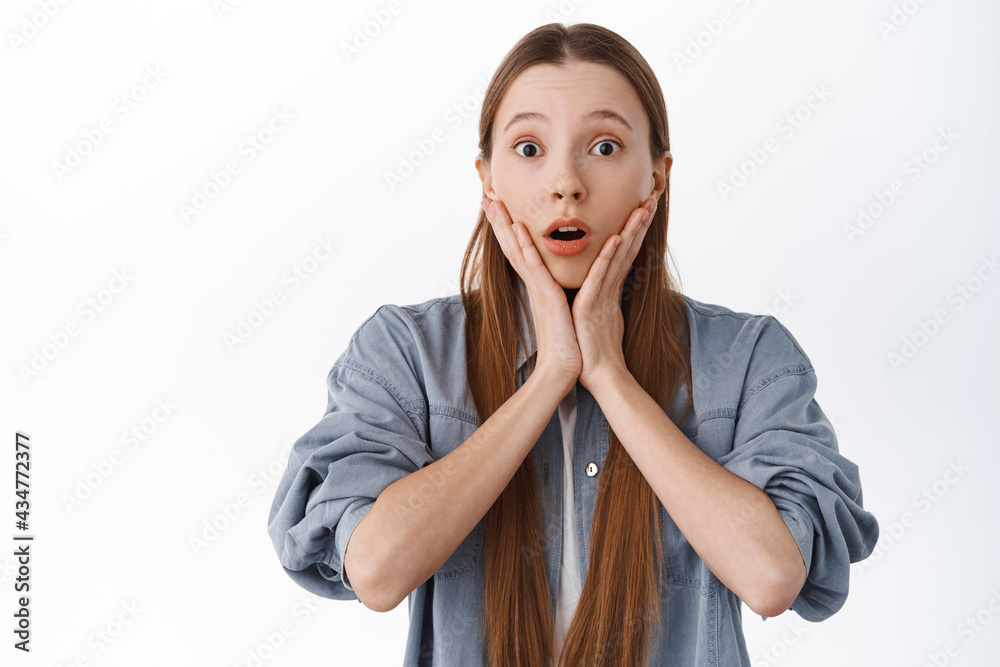 Surprised young woman react to big news, gasping and look in awe, raise eyebrows and open mouth, holding hands on face with shocked expression, white background