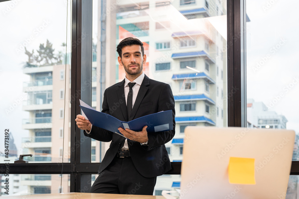 Fototapeta premium Caucasian executive businessman standing holding document folder looking through laptop in the office