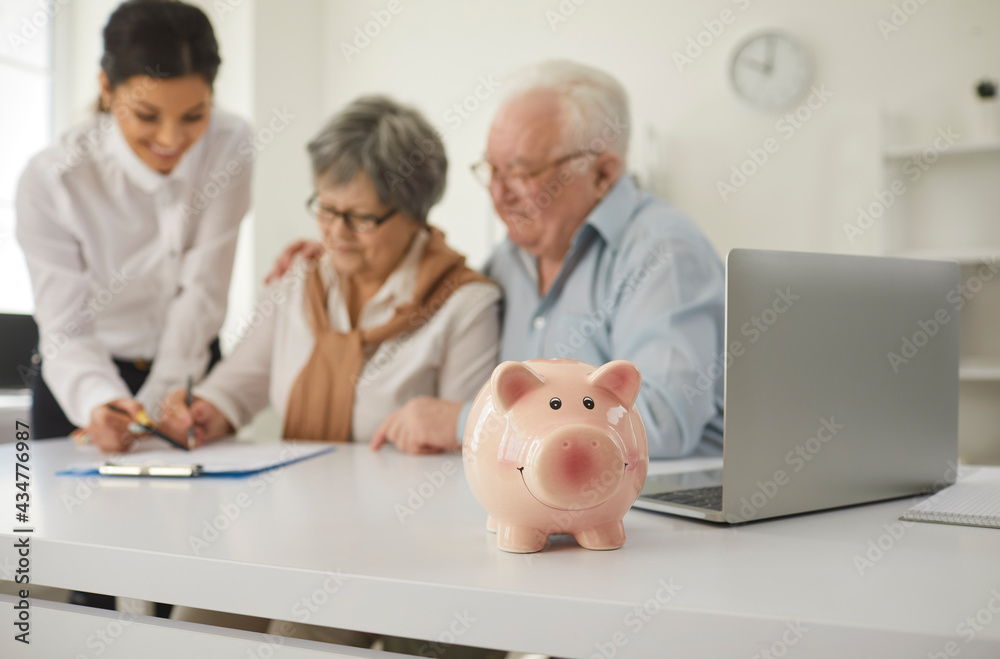 Happy retired senior couple sign life insurance paper at table in broker's office. Older people put signature on money loan agreement sitting at desk with pink piggy bank. Family finance concept Happy retired senior couple sign life insurance paper at table in broker's office. Older people put signature on money loan agreement sitting at desk with pink piggy bank. Family finance concept