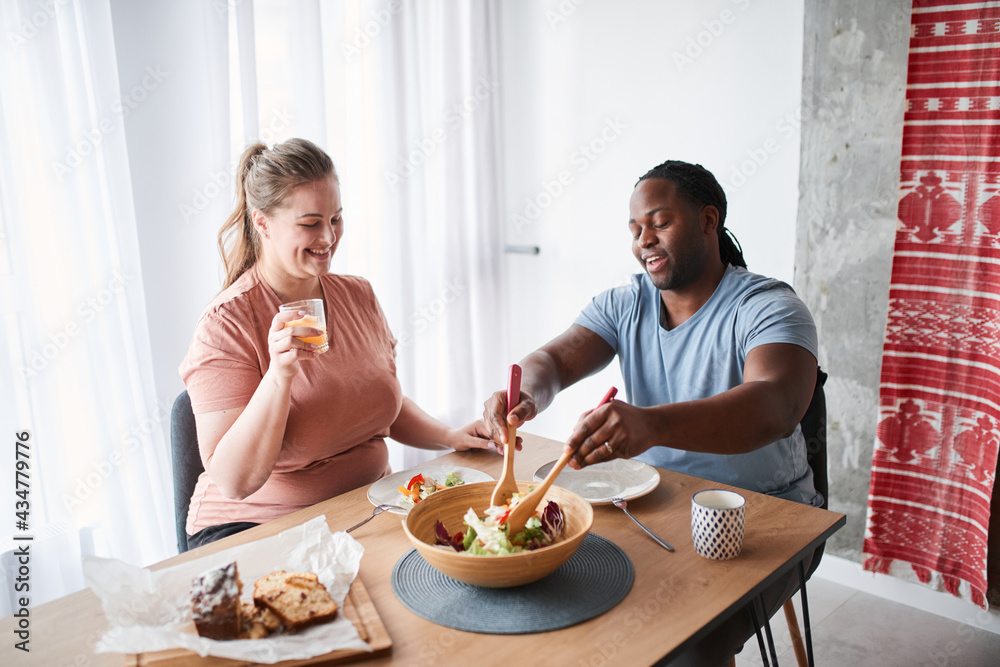 Man mixing salad with wooden spoons while sitting at the table with his wife