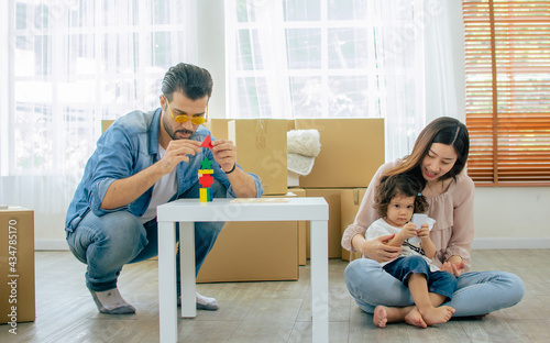 Caucasian handsome father, asian beautiful mother, little mixed race adorable girl or daughter sitting on floor, family playing together with happiness, moving house or apartment with paper boxes.