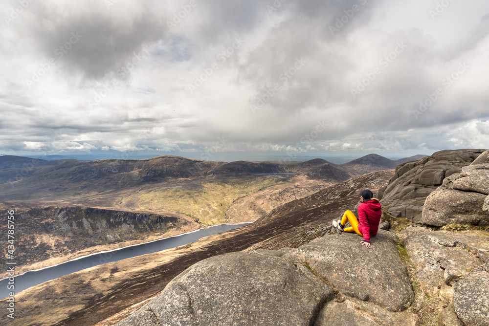 Woman wearing cap, pink jacket, yellow leggings sitting on top of mountain, with lake in background at Slieve Binnian Mountain North Ireland