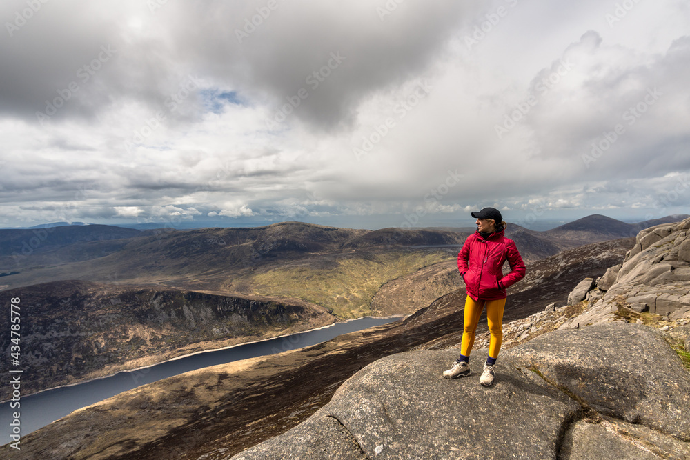 woman wearing cap, pink jacket, yellow leggings standing on top of mountain, with lake in background at Slieve Binnian Mountain North Ireland