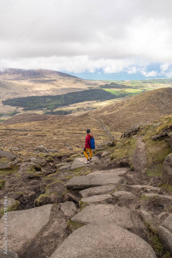 Woman with hat, pink jacket, blue backpack, standing on stones hiking, in the background the Irish Sea and green forests in the Mourne Mountains Newry North Ireland