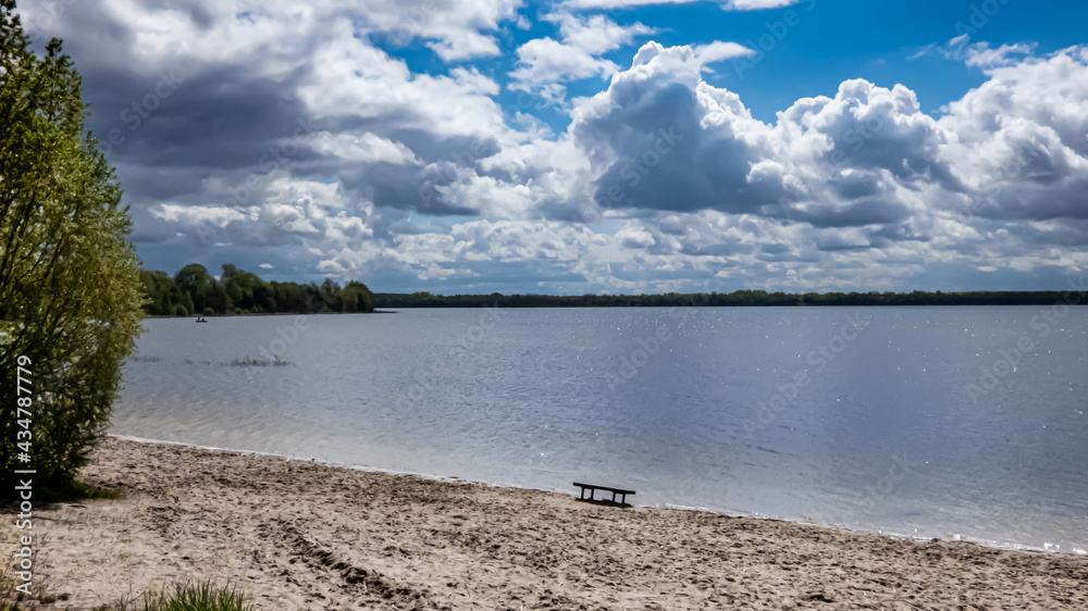 weitwinklige Aufnahme des Dreiweibener See mit wolkig blauem  Himmel und eine Bank am Strand