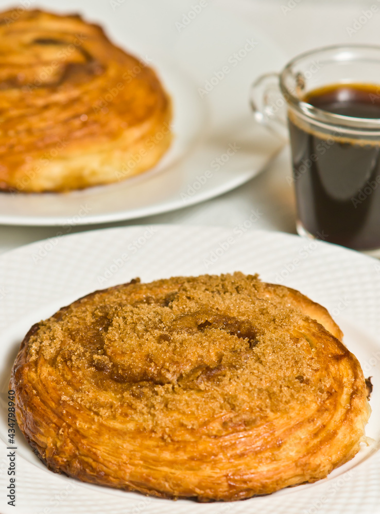 Top, front view, close distance of a freshly baked, croissant cinnamon role on a white, round plate, with a glass cup of espresso, in a red and white stripped place mat