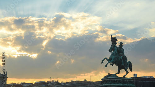 A stone sculpted statue with a horse rider waving a flag above the buildings in Vienna. Sunset, the sun's last rays shine beyond the clouds.
