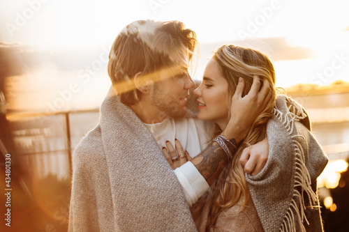 Portrait of happy beautiful couple at the sunset, cuddling in cozy blanket. Charming loving blond man hugging attractive woman, enjoying tenderness, spend time together, romantic date concept