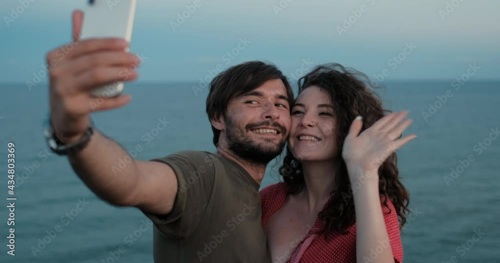 Man and Woman Making Selfie on Smartphone, Kissing and Waving Hands During Video Call, Standing on Hill Against Sea Background in Evening after Sunset Man and Woman Making Selfie on Smartphone, Kissing and Waving Hands During Video Call, Standing on Hill Against Sea Background in Evening after Sunset