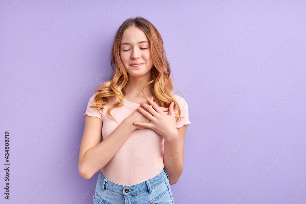 Adorable girl in casual clothes stand holding hands on chest, feeling gratitude or dreaming with eyes closed, calm and dreamy, isolated on purple background, portrait