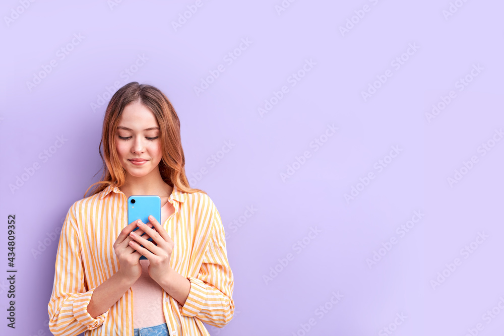 Portrait Of Cute Teenage Girl In Casual Shirt Using Mobile Phone, Chatting, Typing Message To Someone, Smiling, Isolated On Purple Background Portrait Of Cute Teenage Girl In Casual Shirt Using Mobile Phone, Chatting, Typing Message To Someone, Smiling, Isolated On Purple Background