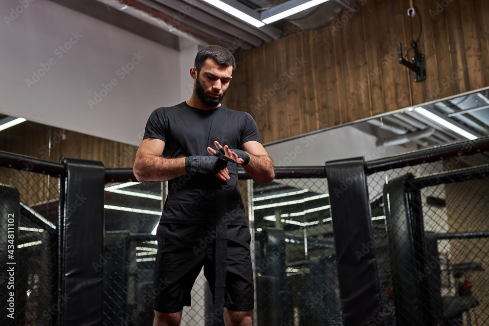 man in black sportswear preparing for tough fight, wrapping fist in sport protective bandages. standing on ring getting ready for mma fight