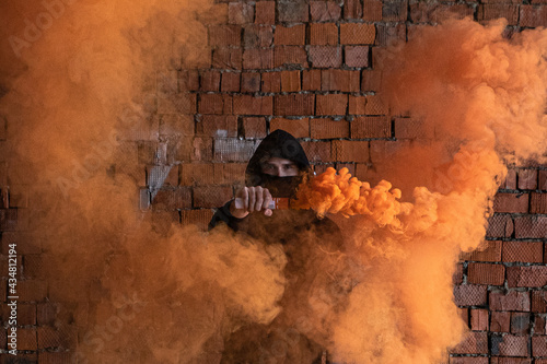 Young handsome criminal model man in a black hoodie with orange fire. Orange smoke bonfire. Hot and explosion, burning gas, danger, and inferno. Black mask, protest of the person