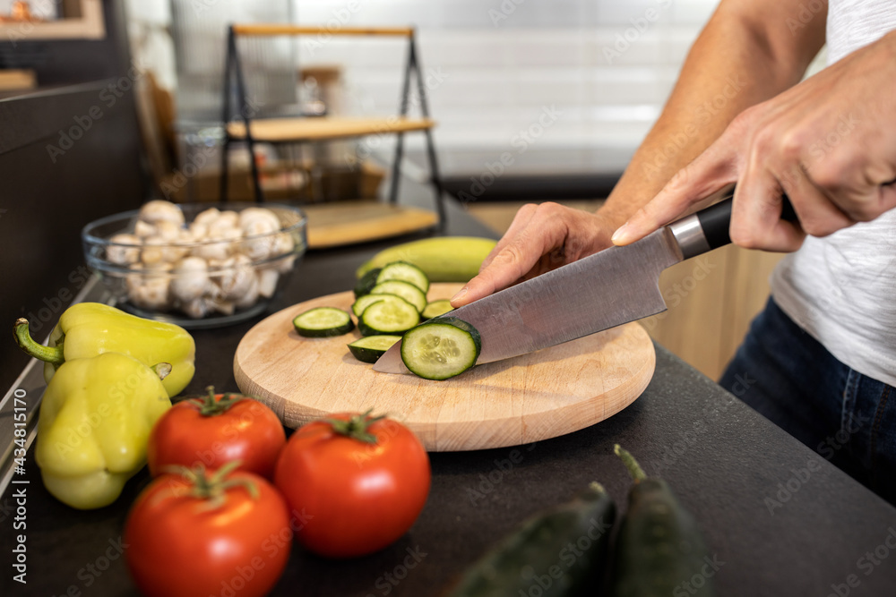 Handsome guy cutting a cucumber with a knife in the kitchen as he prepares a vegetable dish