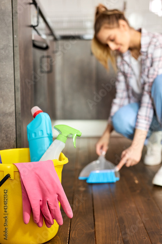 Focus on bucket with pink rubber gloves and detergents inside of it, female housewife removing dust and dirt from floor using brush and dustpan, cleaning house.