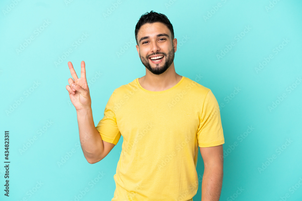 Young arab man isolated on blue background smiling and showing victory sign - obrazy, fototapety, plakaty 