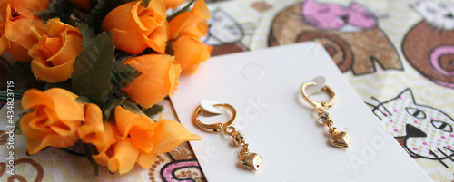 Par de brincos dourados pequenos com argola e pendente com coração no papel branco com tecido estampado e flores laranjas ao fundo. photo