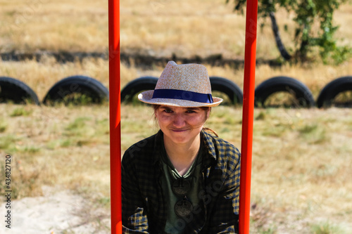 Defocus smiling young woman in checkered shirt and hat swinging on swing on playground. Hipsters and hippie portrait. Countryside area. Bright red swing. Out of focus