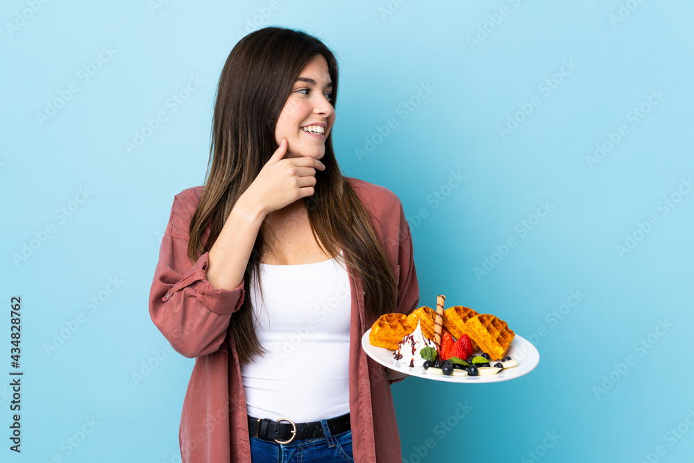 Teenager Brazilian girl holding waffles over isolated blue background thinking an idea and looking side Teenager Brazilian girl holding waffles over isolated blue background thinking an idea and looking side