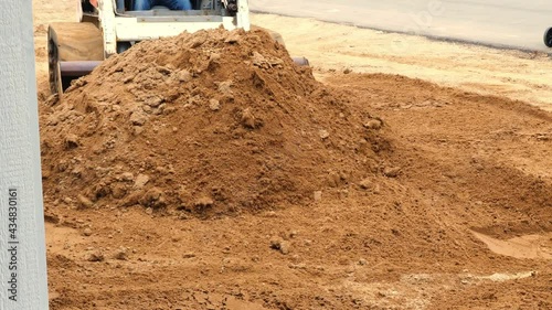Compact front loader pushes a pile of brown dirt to spread it out, as preparation of the landscape before sod is added, at a construction job site.