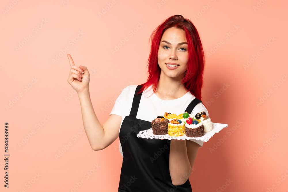 Pastry chef holding a muffins isolated on pink background happy and pointing up Pastry chef holding a muffins isolated on pink background happy and pointing up