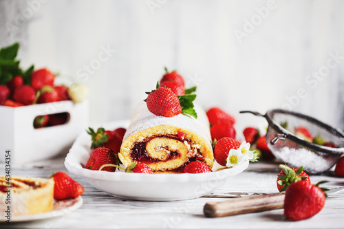 Homemade strawberry shortcake cake roll or Roulade with a berry jam filling and powdered sugar with mint leaves. Dessert over a white rustic table. Selective focus with blurred foreground  background