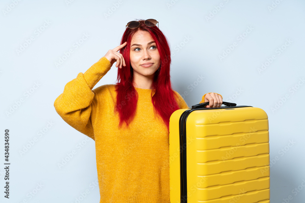 Traveler girl holding a suitcase isolated on blue background having doubts and thinking Traveler girl holding a suitcase isolated on blue background having doubts and thinking