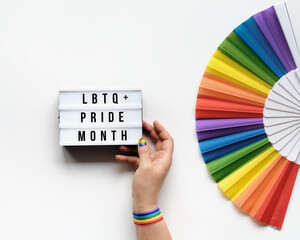 LGBTQ pride month decor. Text Happy Pride Month on lightbox. Rainbow pattern fan on off white background. Human hand with rainbow nail varnish holds light box. June, LGBT pride month pride symbols.