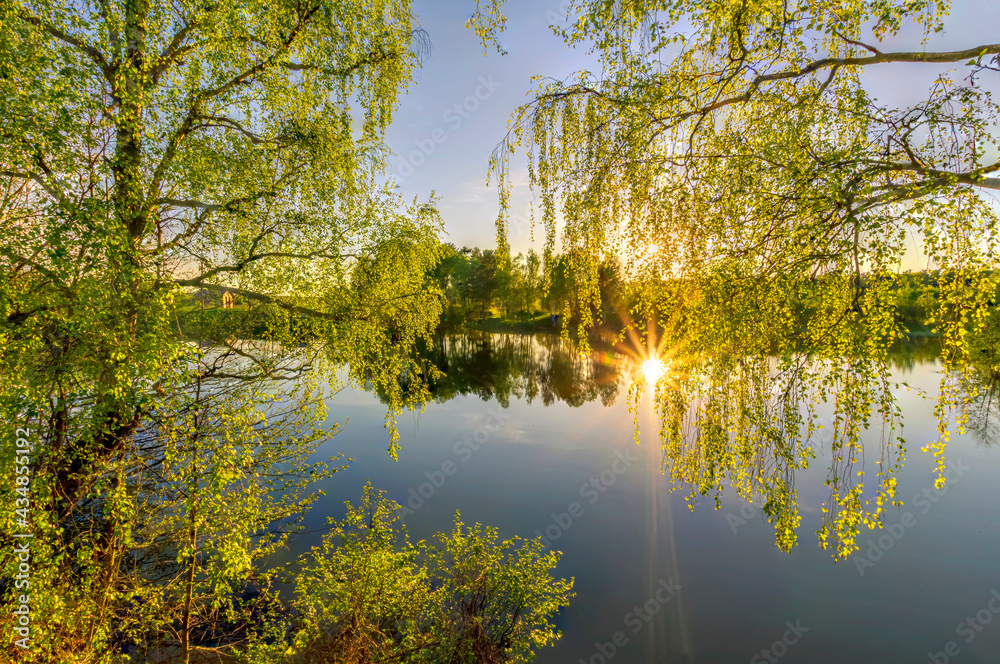 Scenic view at beautiful spring sunset on a shiny lake with green branches, birch trees, bushes, grass, golden sun rays, calm water ,deep blue cloudy sky and forest on a background, spring landscape