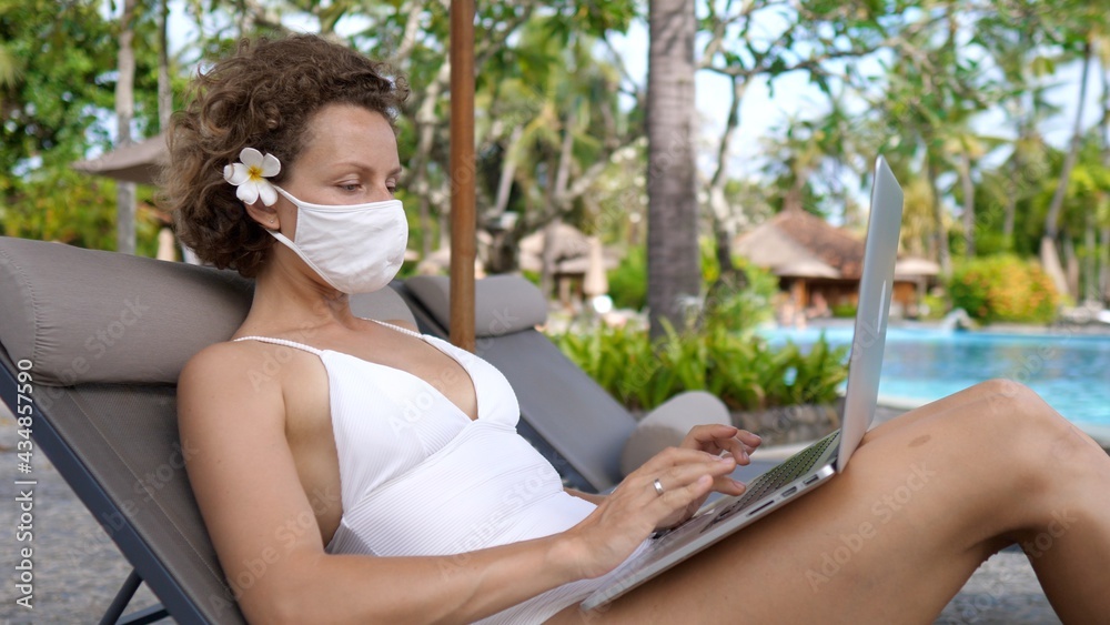 Digital nomad concept. Young beautiful woman in protective mask and a white swimsuit works on a laptop by the pool. 
