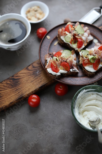 Three toasts with bacon, cherry tomatoes, cucumber and cream cheese. Sandwiches on a ceramic plate on a wooden board on a dark table. Snacks, top view.