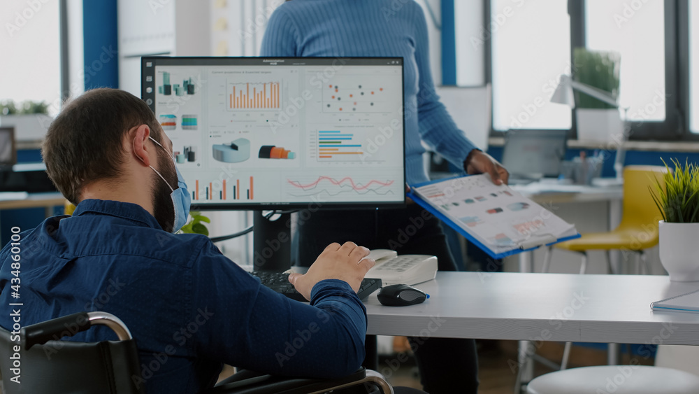 Diverse businesspeople using computer desktop, showing new startup idea, sitting at desk in new normal company office wearing protection mask, team leader with disabilities sitting in wheelchair