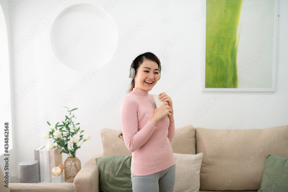 Happy young Asian woman in earphones singing and listening to music and holding mobile phone at home.