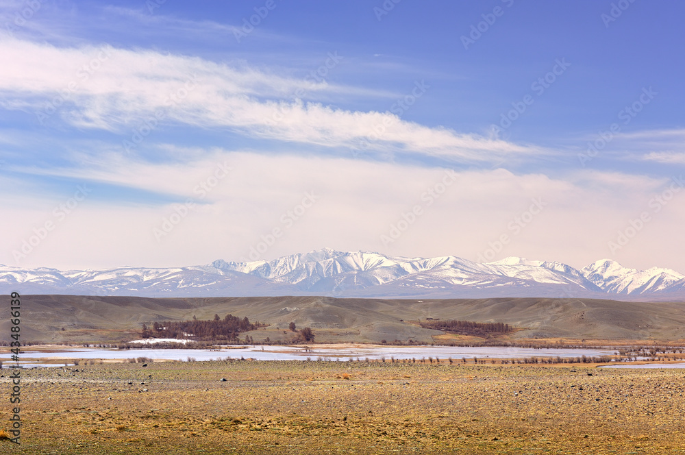 Chui steppe in the Altai Mountains. Lake in the steppe, snow-covered North Chui ridge in the distance under a blue sky with clouds. Pure Nature of Siberia, Russia