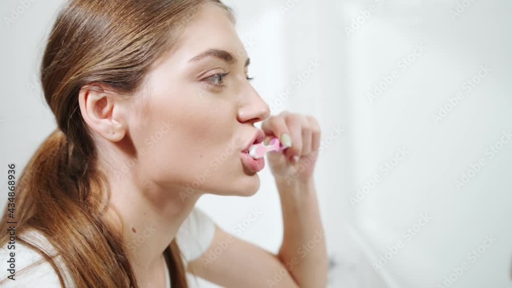Side view portrait of attractive woman looking at mirror and brushing teeth at home or hotel. Concept of doing water procedures with good mood at home. Side view portrait of attractive woman looking at mirror and brushing teeth at home or hotel. Concept of doing water procedures with good mood at home.