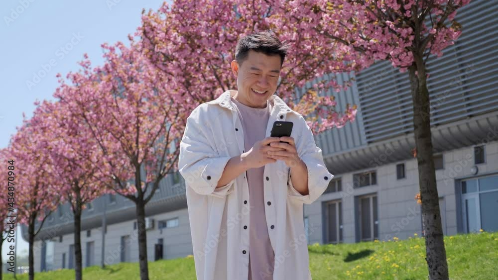 Positive Lifestyle Asian Korean man Chatting on his Smartphone Looking Away Smiling on the background of beautiful rose trees and modern building . Checking News, Mails. Looking Confident, Successful. Positive Lifestyle Asian Korean man Chatting on his Smartphone Looking Away Smiling on the background of beautiful rose trees and modern building . Checking News, Mails. Looking Confident, Successful.
