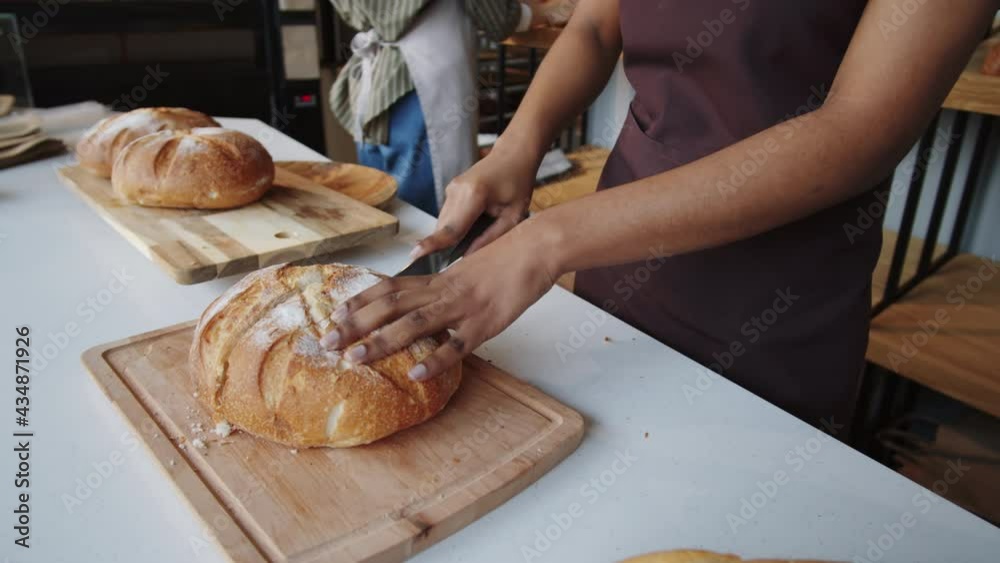 Beautiful Afro-American woman in apron cutting freshly baked loaf of bread with knife while working in bakery Beautiful Afro-American woman in apron cutting freshly baked loaf of bread with knife while working in bakery