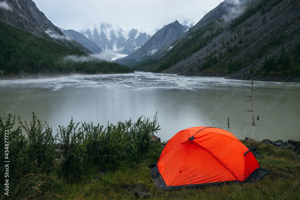Atmospheric alpine landscape with orange tent on shore of green mountain lake and snowy mountains in rainy weather. Gloomy scenery with rainy circles on water of mountain lake and low clouds in valley