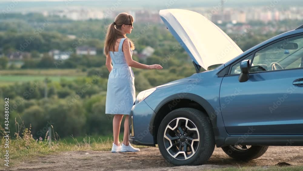Young female driver standing alone near a broken car with popped up hood inspecting her vehicle engine and waiting for help. Young female driver standing alone near a broken car with popped up hood inspecting her vehicle engine and waiting for help.
